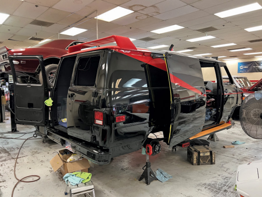 A side view of a black van in a garage with its doors open. The van has a distinctive red spoiler on the roof and a red stripe running diagonally down its side. The van is lifted on jacks, with its rear wheel removed, exposing the brake assembly. In the background, there is another red car on a lift and a large fan to the right. Tools and various parts are scattered on the floor around the van.