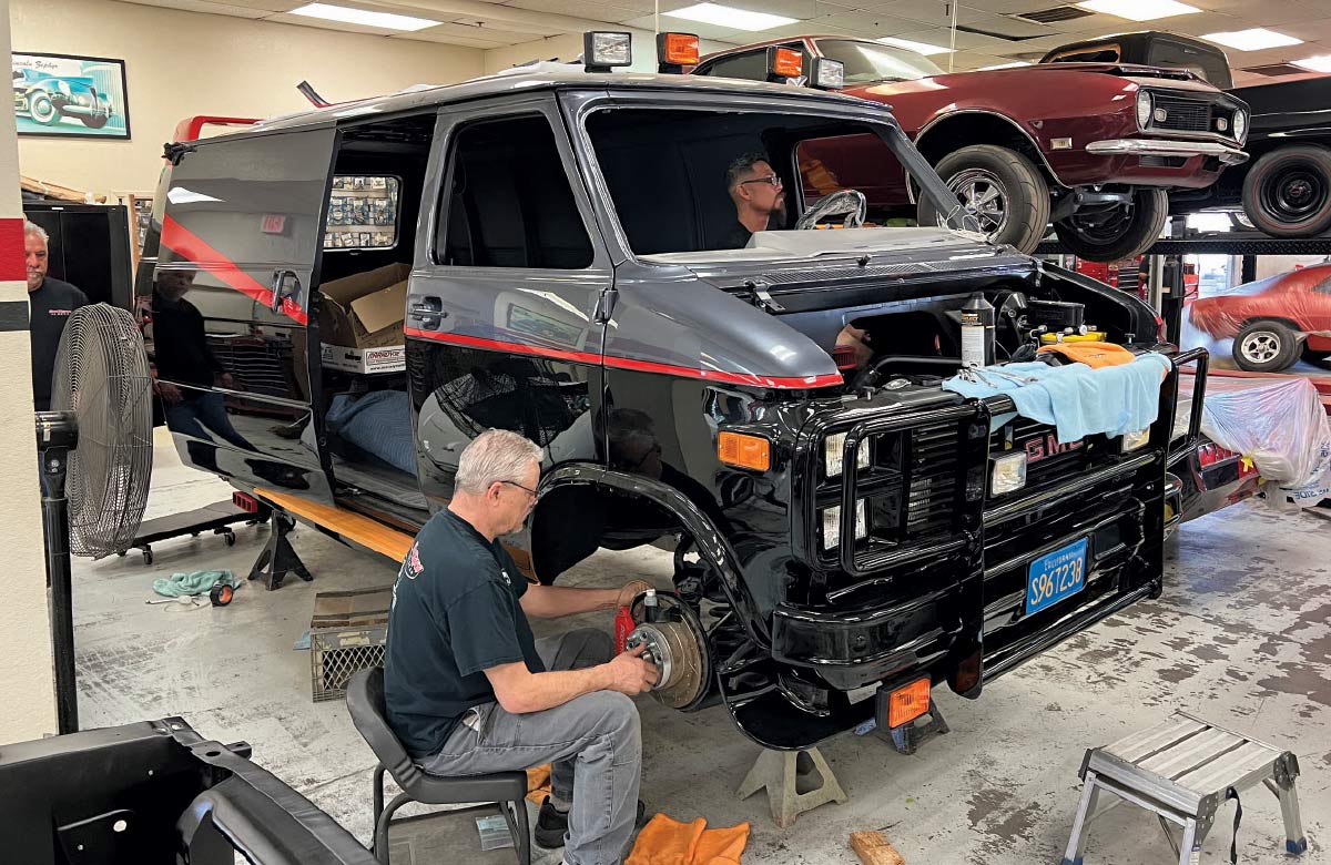 A group of men are working on a black van inside a garage. The van is jacked up and missing its front wheels. One man in a black T-shirt is sitting on a stool, working on the front brake assembly. Another man is sitting in the driver's seat of the van. The van has a red stripe running along its side and a roof rack with spotlights. In the background, there is a red muscle car on a lift. A fan and various tools are scattered on the floor.