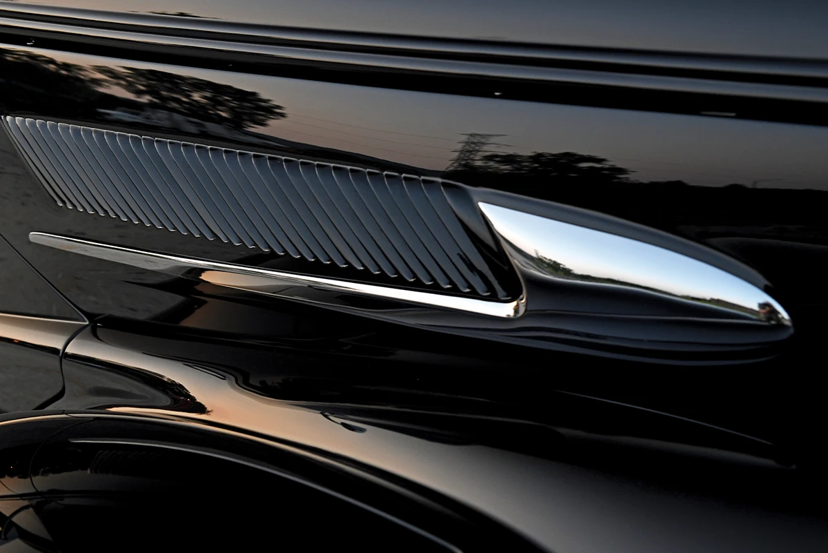 Close-up of a custom black hot rod's glossy fender and integrated chrome side vent detail, showing reflections of the surrounding area