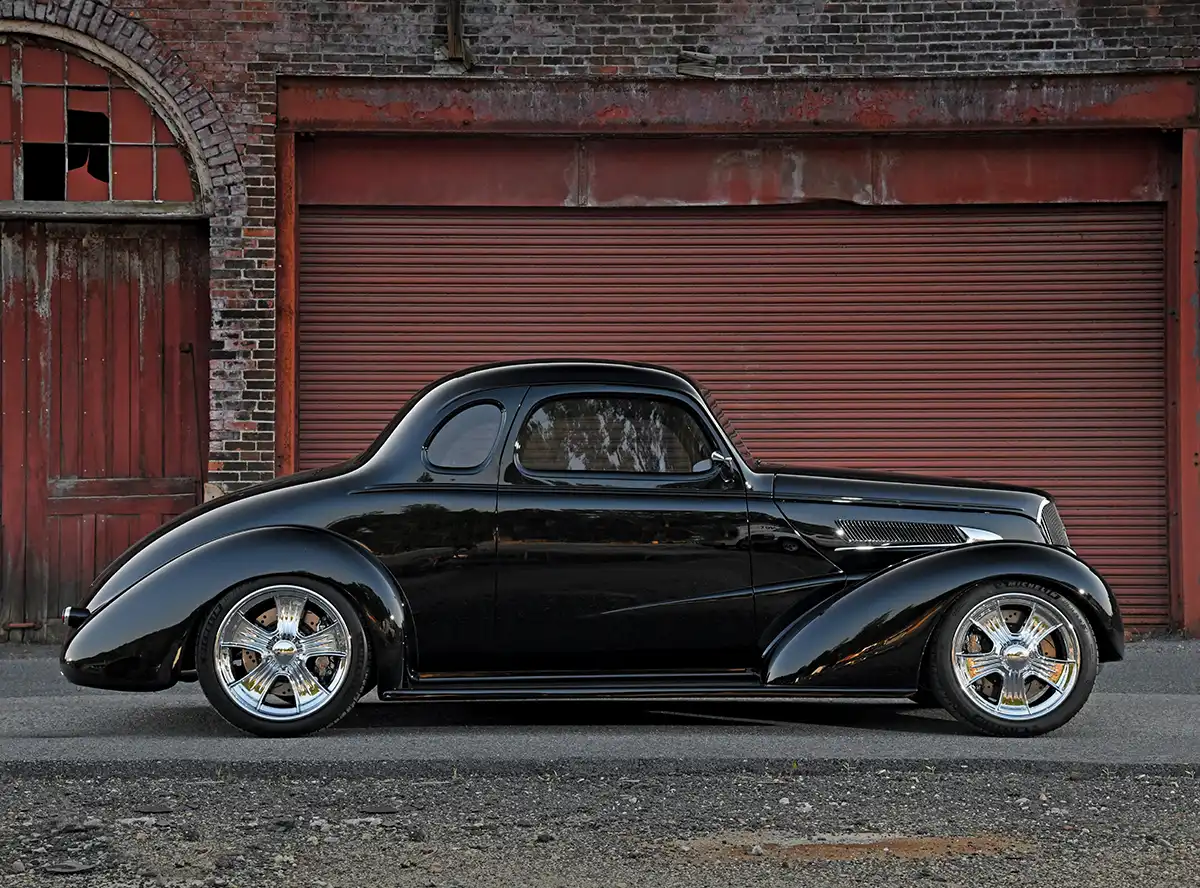 Profile view of a custom, high-gloss black 1930s coupe hot rod with chrome wheels parked in front of a red brick building and garage door