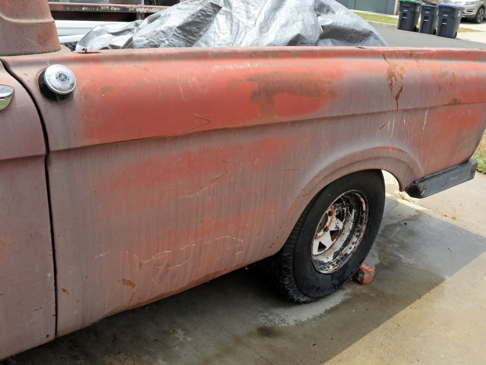 Side view of an old pickup truck's rusty, reddish-brown rear quarter panel. The paint is faded, and rust is visible around the wheel well and fuel filler cap.