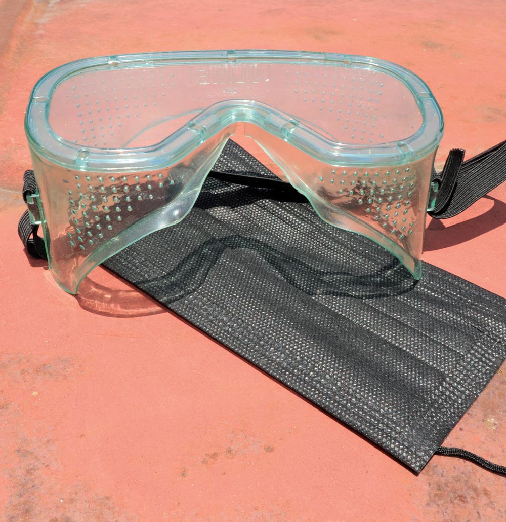 Safety gear, including a pair of clear plastic goggles with a perforated edge and a black disposable face mask, resting on a reddish, rusty metal surface.