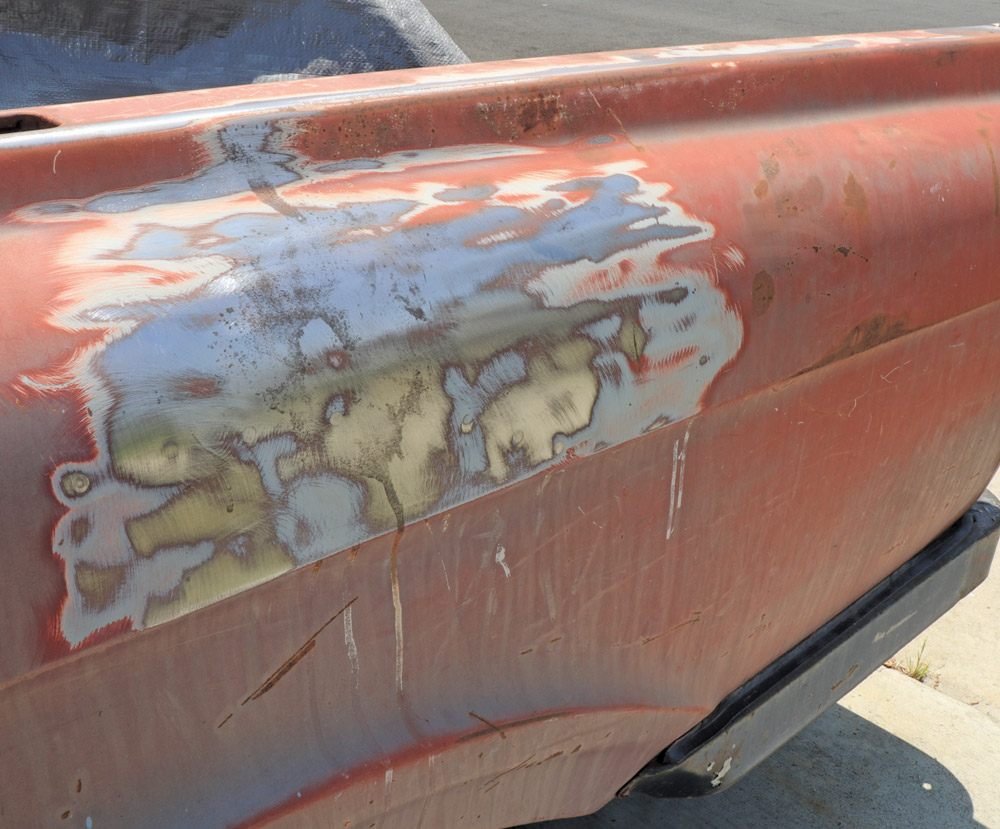 The side panel of a truck with large sections of paint removed, exposing bare metal and patchy remnants of body filler or primer, indicating prep work for restoration.