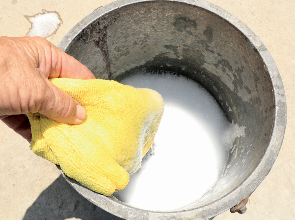 A hand holding a wet, yellow microfiber cloth dipping into a metal bucket filled with soapy, white water, likely for cleaning the vehicle surface.