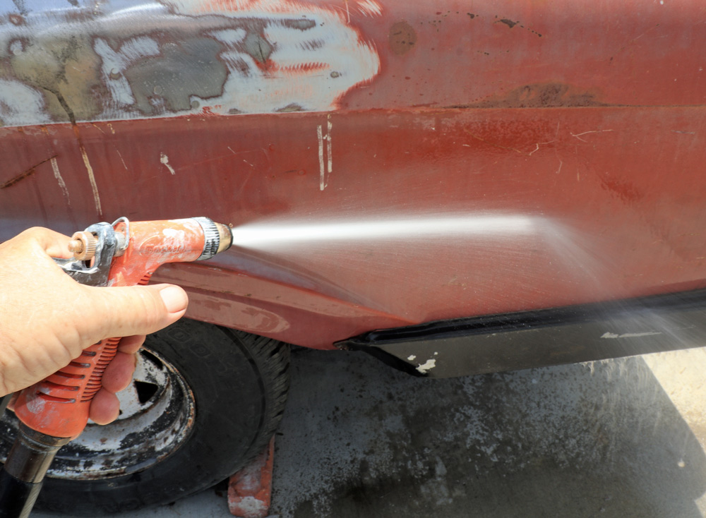 A hand holding an orange spray nozzle, rinsing a section of the truck's side panel with a stream of water after sanding or cleaning.