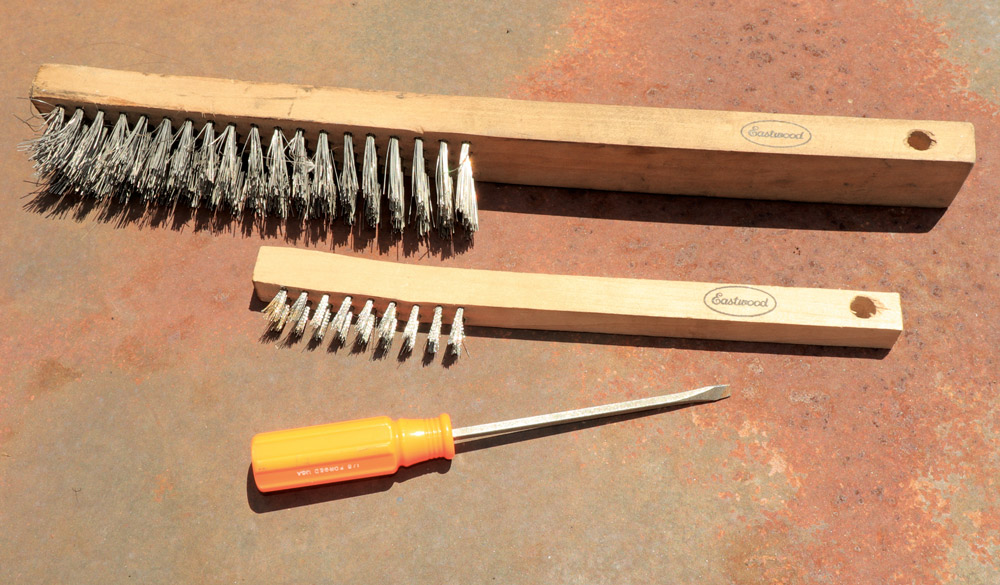 Two wooden-handled wire brushes of different sizes, and a small, orange-handled flathead screwdriver, resting on a rusty, reddish metal surface.