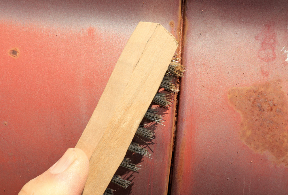 A hand uses a small, wooden-handled wire brush to aggressively scrub rust and residue out of a seam where two panels of the truck body meet.