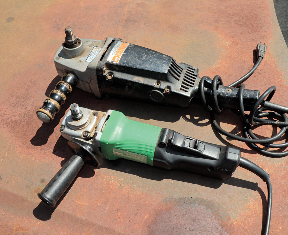 Two angle grinders resting on a rusty red surface: a larger, older one with a handle attached, and a newer, smaller green and black one.