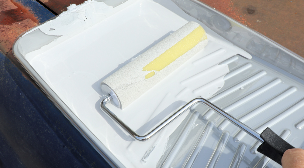 A paint roller loaded with white primer resting in a disposable paint tray filled with the mixed paint material.