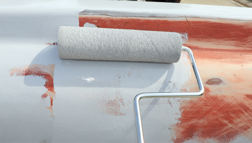 A close-up of a paint roller applying a thick coat of white primer to the top rail of the truck bed, covering the exposed red and bare metal.