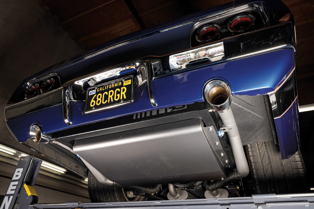 Undercarriage view from the rear, showing the custom exhaust system exiting under the chrome bumper, a large fuel tank/sump, and the rear suspension components.