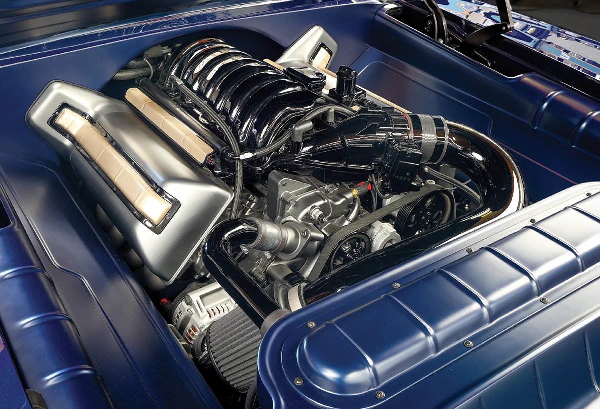 Engine bay view showing a modern, high-performance engine in a smooth, custom-painted blue bay. The engine has polished silver intake manifolds and a black ribbed cover.