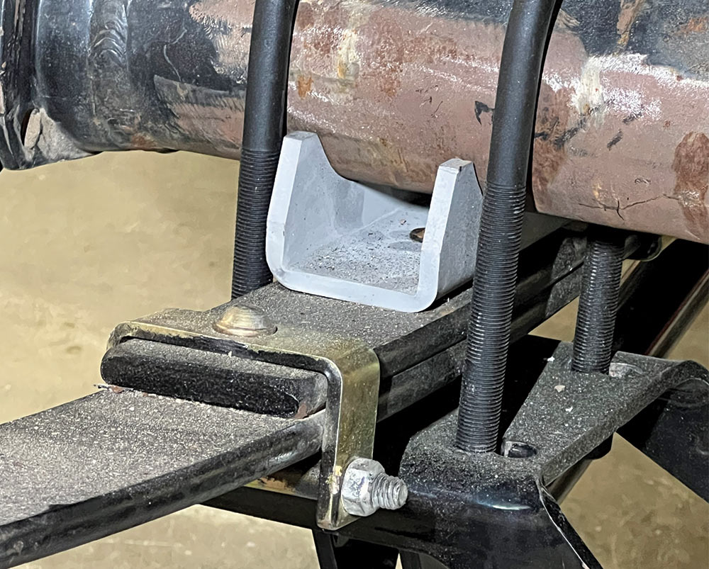 Close-up of a vehicle's suspension showing a gray metal spacer block resting on a leaf spring assembly secured by two large U-bolts.