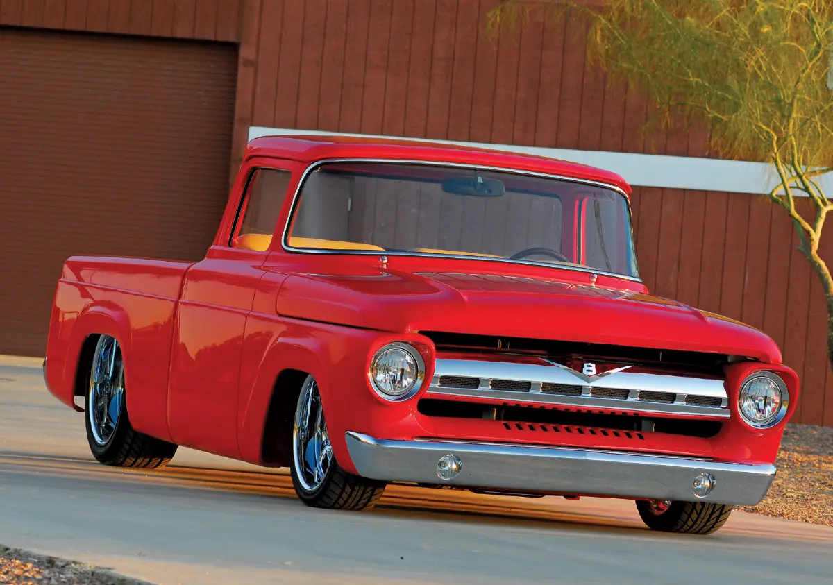 Bright red, customized Ford vintage hot-rod style pickup truck parked on a light-colored paved driveway; In the background, there is a large red barn and a thin, leafless tree under a soft, golden-hour light