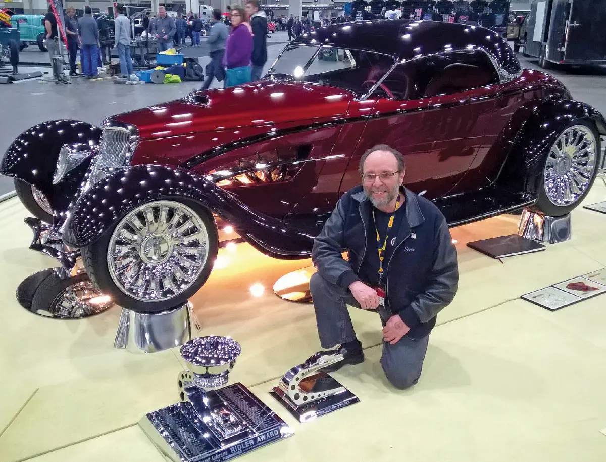 Steve Frisbie smiling in prescription eyeglasses, a black jacket, and dark grey pants as he took home the 2017 Ridler award with Buddy Jordan’s highly modified deep-red vintage 1933 Ford roadster with large chrome wheels as he is crouched kneeling down next to the award and some other chrome award next to that plus the roadster behind him inside a car show setting
