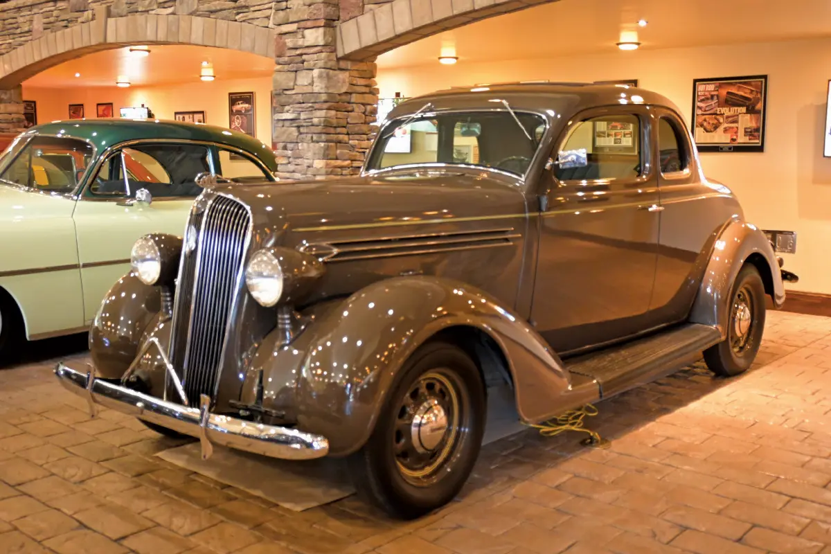 A polished brown 1936 Plymouth vintage coupe parked inside a showroom with a stone-paved floor and arched stone pillars; Other classic cars are visible in the background, along with a wall-mounted monitor displaying an image of a car