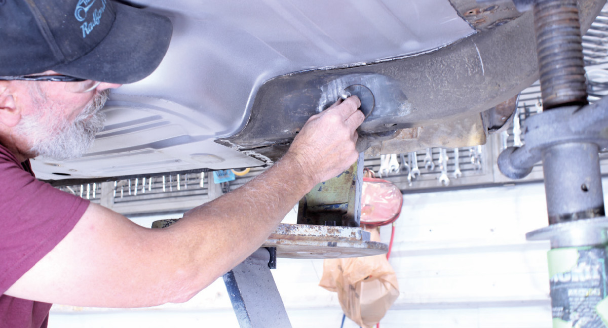 A mechanic in a baseball cap working on the undercarriage of a vehicle, adjusting a mounting plate on the frame.