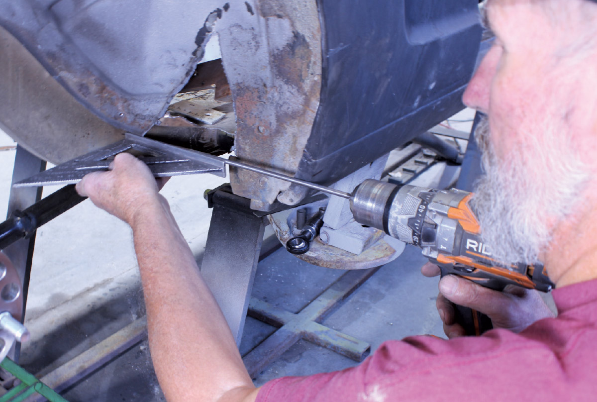 A mechanic uses a power drill on a vehicle frame while checking alignment with a metal speed square.