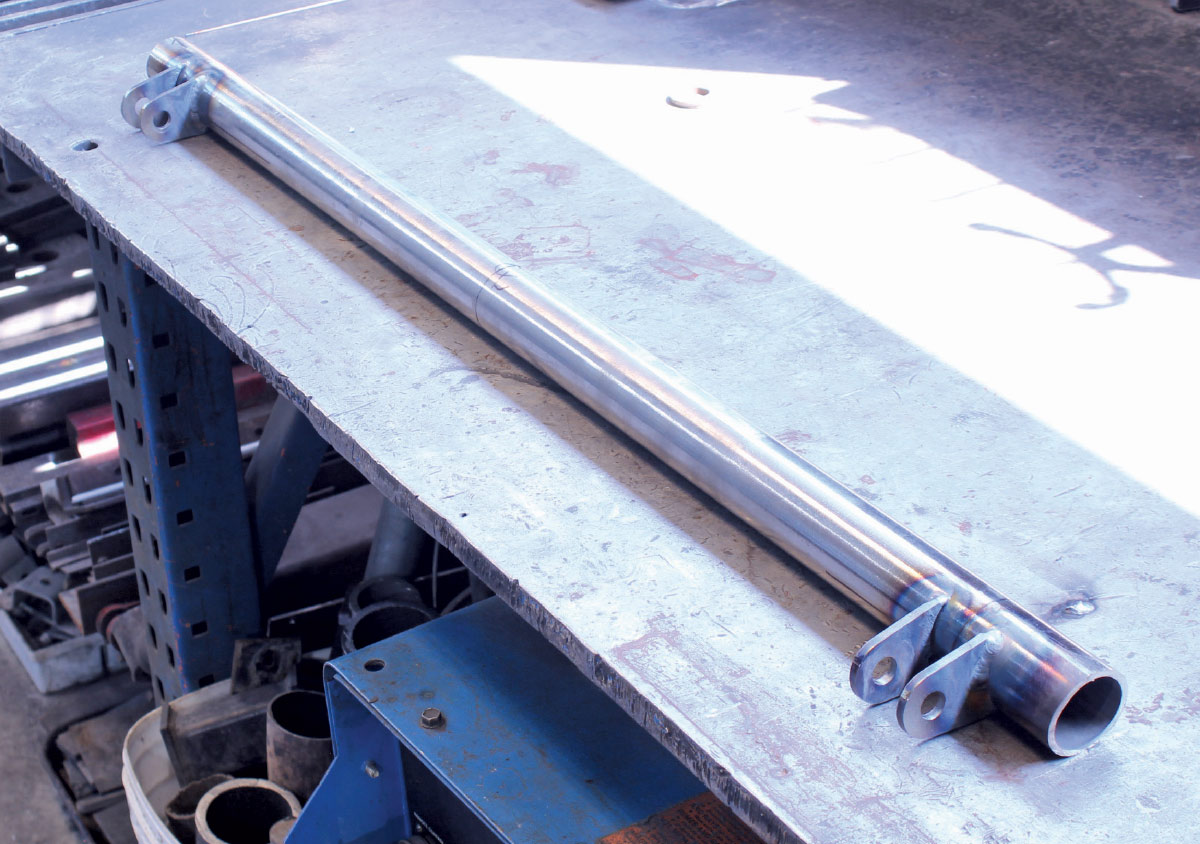 A long, silver machined suspension tube with welded mounting brackets at both ends resting on a metal workbench.