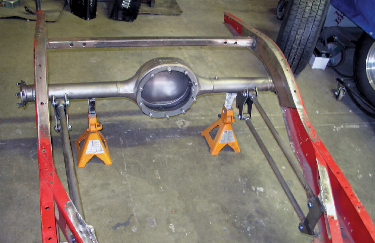 A bare vehicle frame on jack stands with a rounded rear axle housing and long suspension links positioned for fitting.