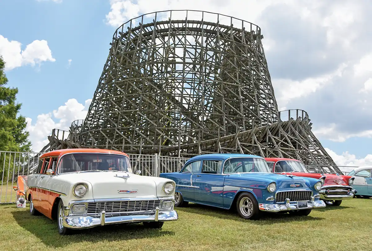 Four classic Chevrolet Tri-Five cars parked on grass, wooden roller coaster structure background