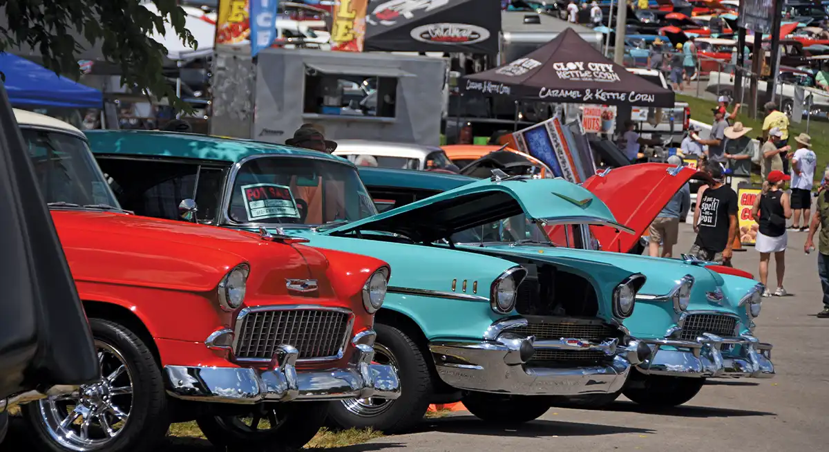 Red and turquoise 1955 Chevrolets parked side by side, vendor tents, car show atmosphere