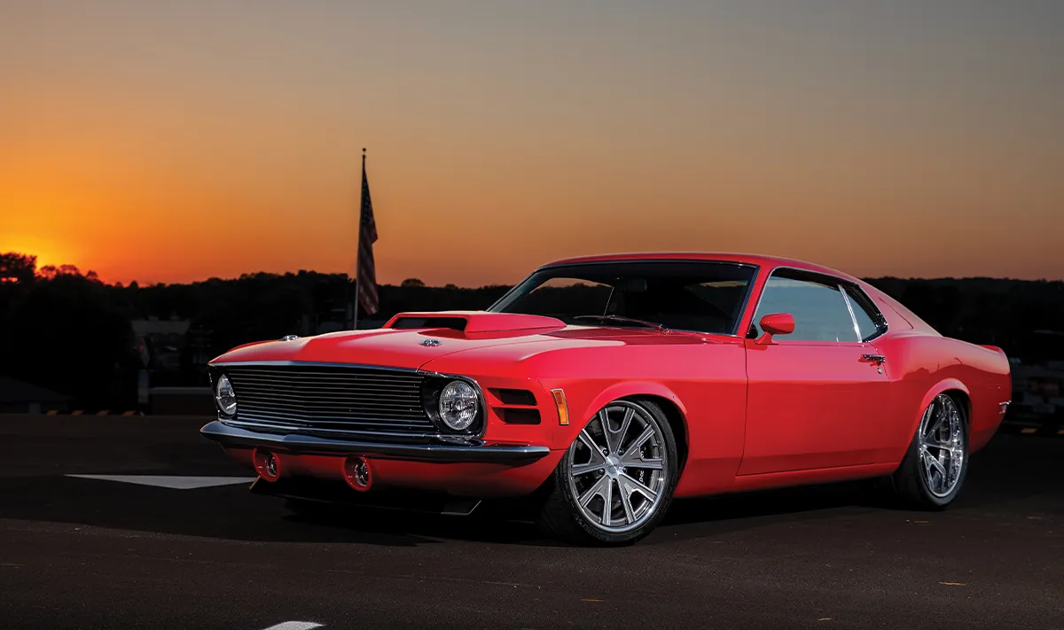 A red 1970 Ford Mustang restomod with custom silver wheels parked on an asphalt lot during a vibrant sunset.