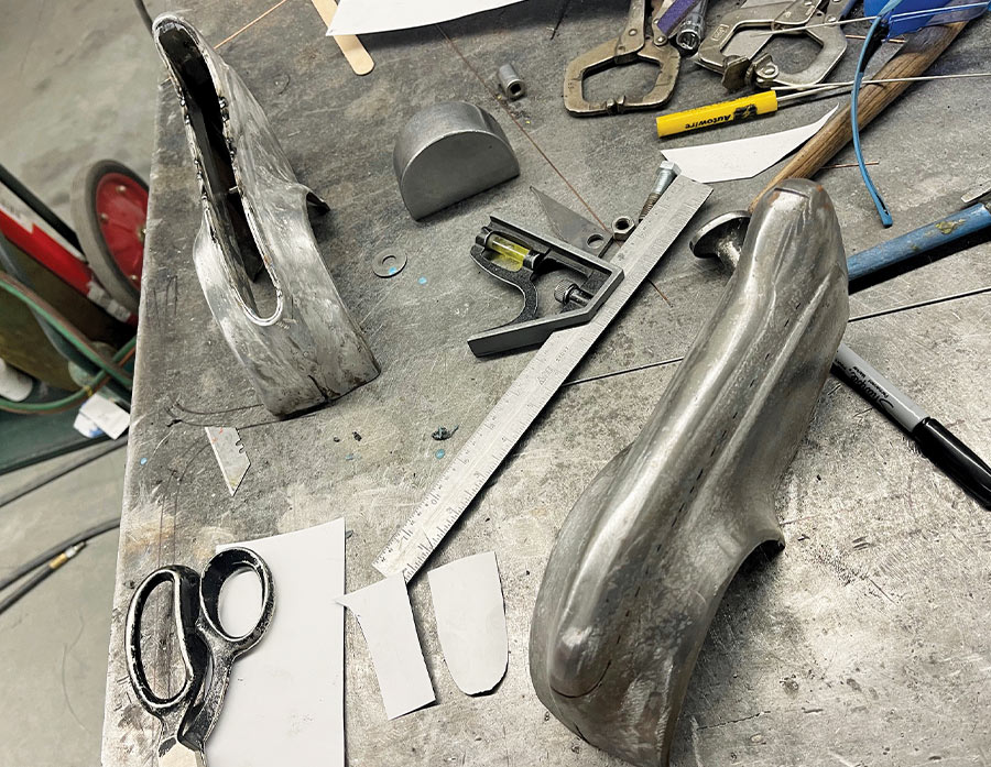 Metal fabrication workbench featuring hand-formed aluminum components, a combination square, snips, and various measuring tools.