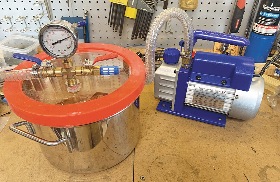 A stainless steel vacuum degasification chamber and a blue electric vacuum pump set up on a workshop table for resin casting.