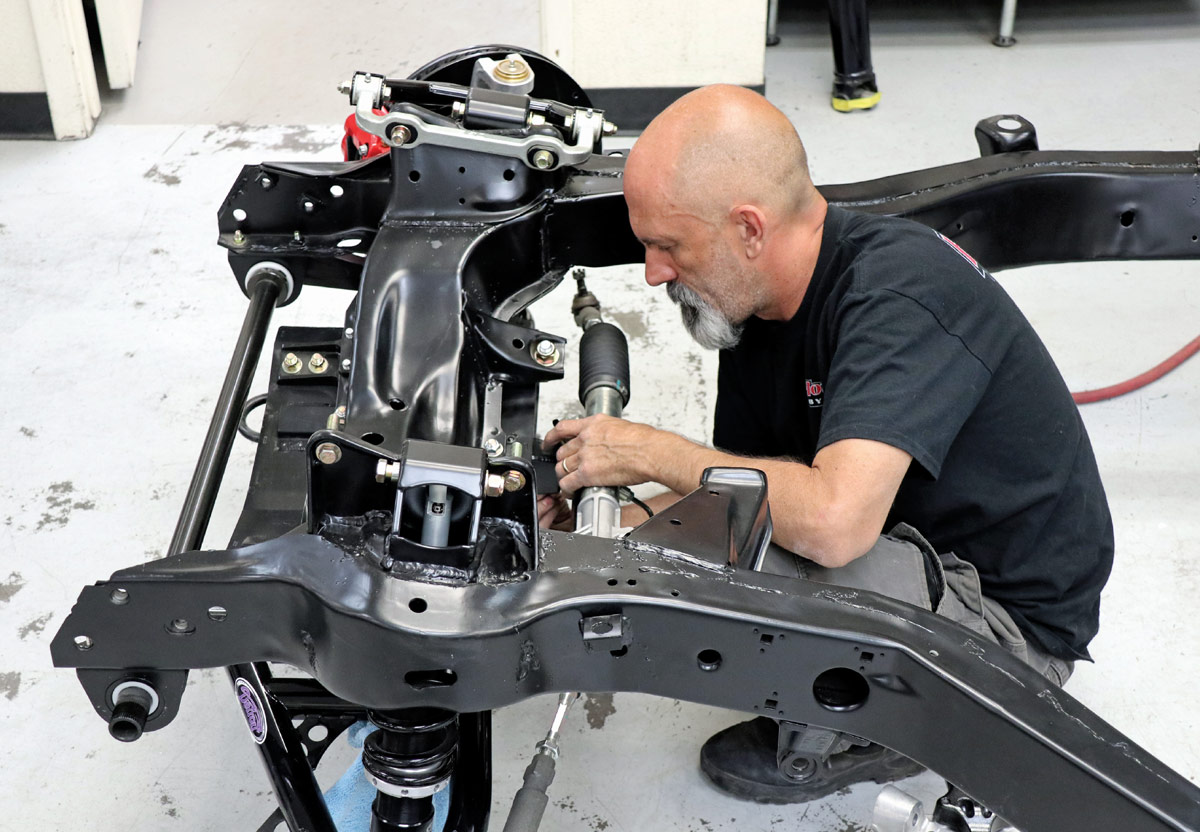 A technician installs a new steering rack into a black powder-coated Corvette frame, carefully aligning mechanical hardware within a clean, professional shop environment.