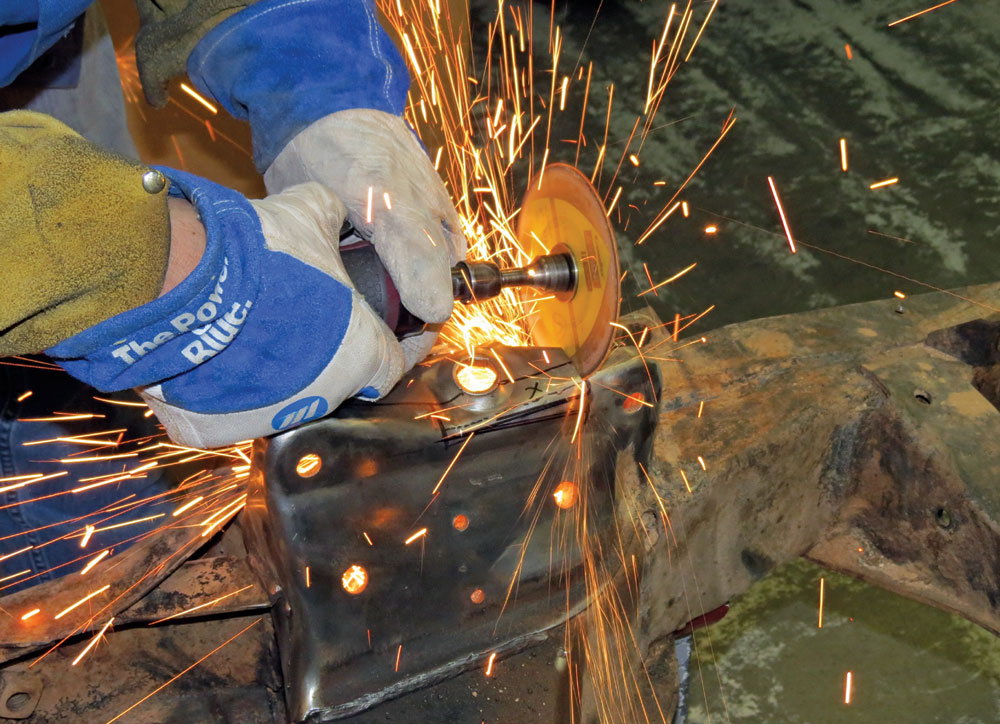 A worker wearing protective gloves uses a power grinder to cut into the rusted Corvette frame, creating bright orange sparks as they prep the metal for new components.