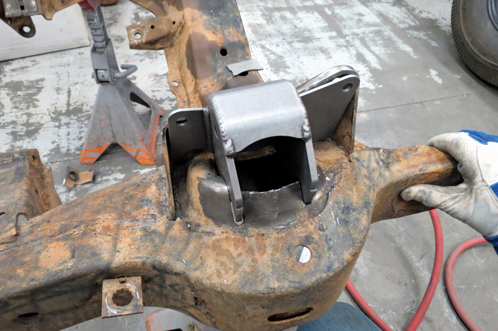A new, unpainted steel suspension bracket is test-fitted onto the original rusted Corvette frame, showing the contrast between the old chassis and new performance parts.