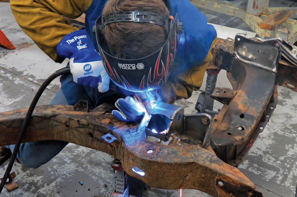 A technician wearing a welding helmet and protective gear uses a MIG welder to permanently attach new steel brackets to the original Corvette frame in a shower of blue light.