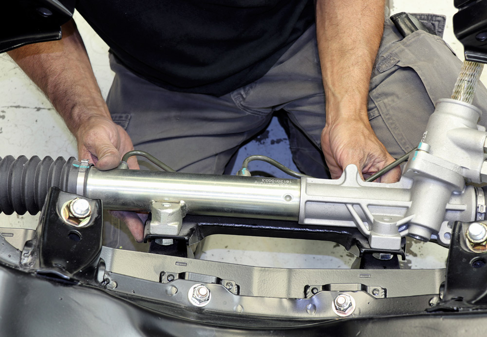 Close-up of a technician's hands positioning a polished silver steering rack onto the black powder-coated chassis, securing the unit with heavy-duty mounting hardware.