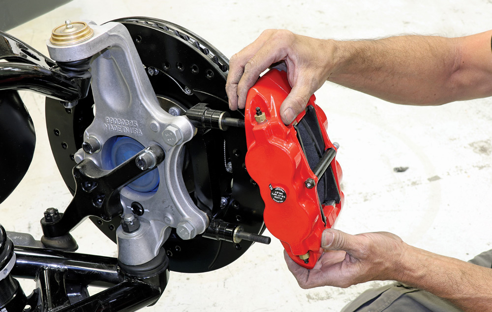 A technician's hands carefully slide a bright red, six-piston Wilwood brake caliper onto the mounting studs of a newly assembled front suspension with a black slotted rotor.