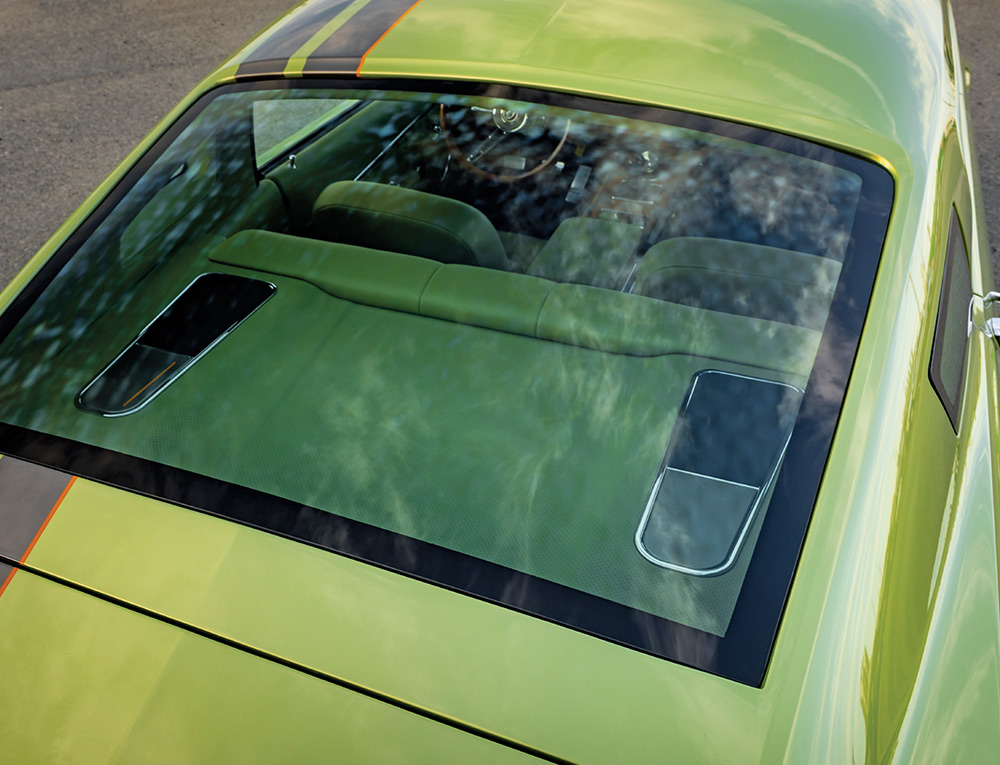 A high-angle view looking through the large fastback rear window of the lime green car, showing the custom interior, rear deck speakers, and the black racing stripes on the roof.