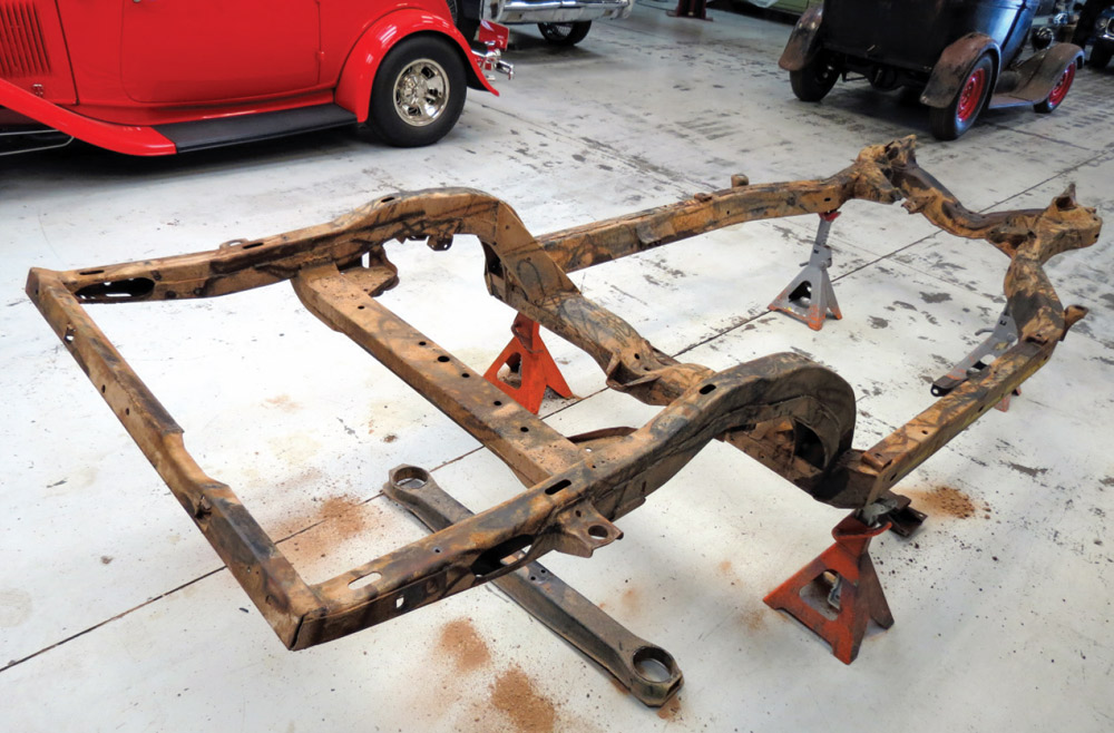 A bare, rusted 1971 Corvette chassis frame sits on orange jack stands in a shop. It has been stripped of its rear crossmember, bumper brackets, and various mounts in preparation for the new Detroit Speed suspension installation.