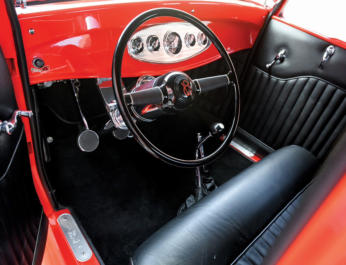 Interior of a red 1931 Ford hot rod featuring black tuck-and-roll upholstery. The red dashboard has an engine-turned gauge panel. Includes a black steering wheel with a red V8 logo, a floor shifter, and chrome hardware.