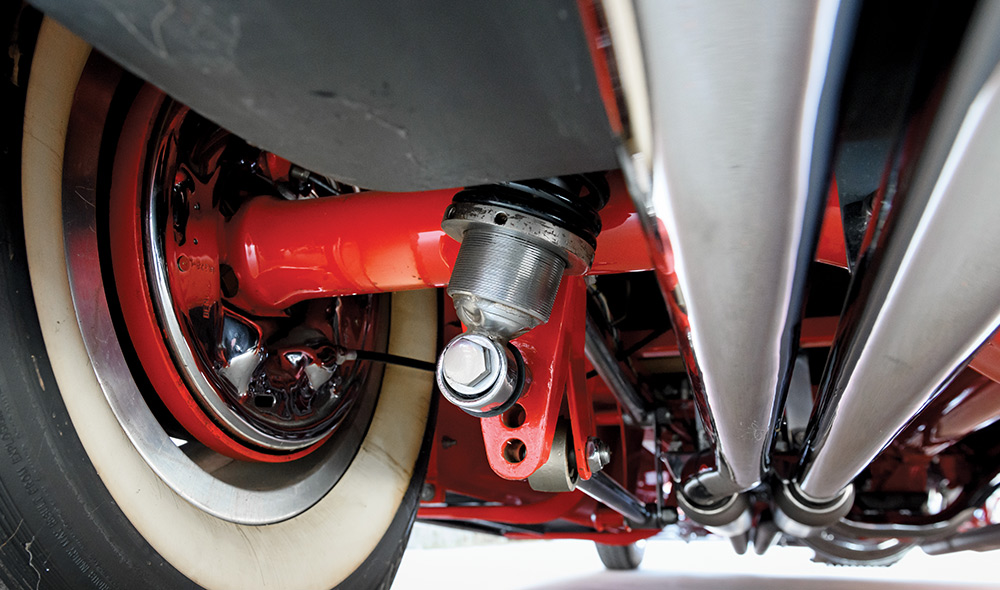 Close-up of the rear suspension under a red '31 Ford. Features a red painted axle, chrome coilover shock, and polished dual exhaust pipes. A wide whitewall tire with a chrome rim is visible on the left.