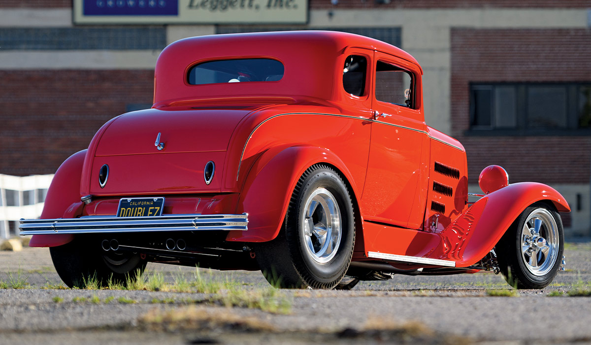 Rear three-quarter view of the red chopped '31 Ford Coupe. Highlights the smooth decklid, oval taillights, and California license plate "DOUBLE Z." Features wide rear tires on chrome wheels and a horizontal triple-bar chrome bumper.