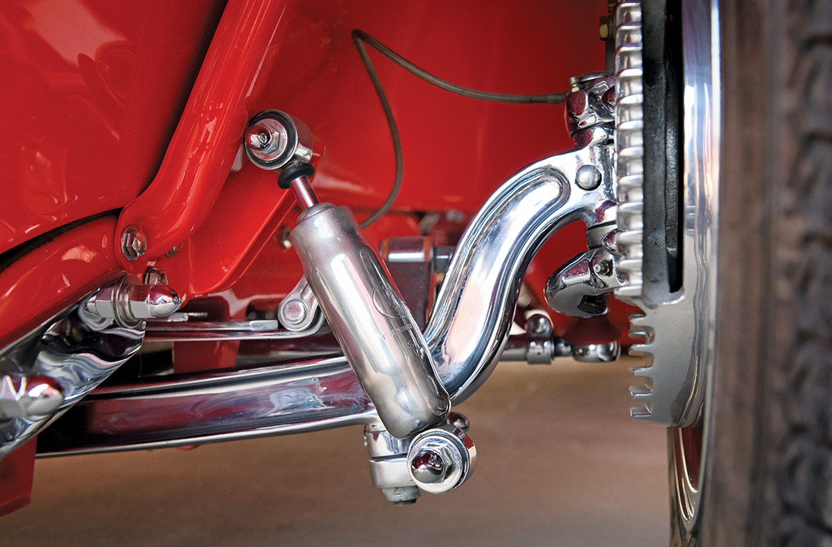 Extreme close-up of a front suspension showing a chrome-plated dropped axle and steering linkage. A polished shock absorber is mounted at an angle next to a finned brake drum and a chrome wheel rim.