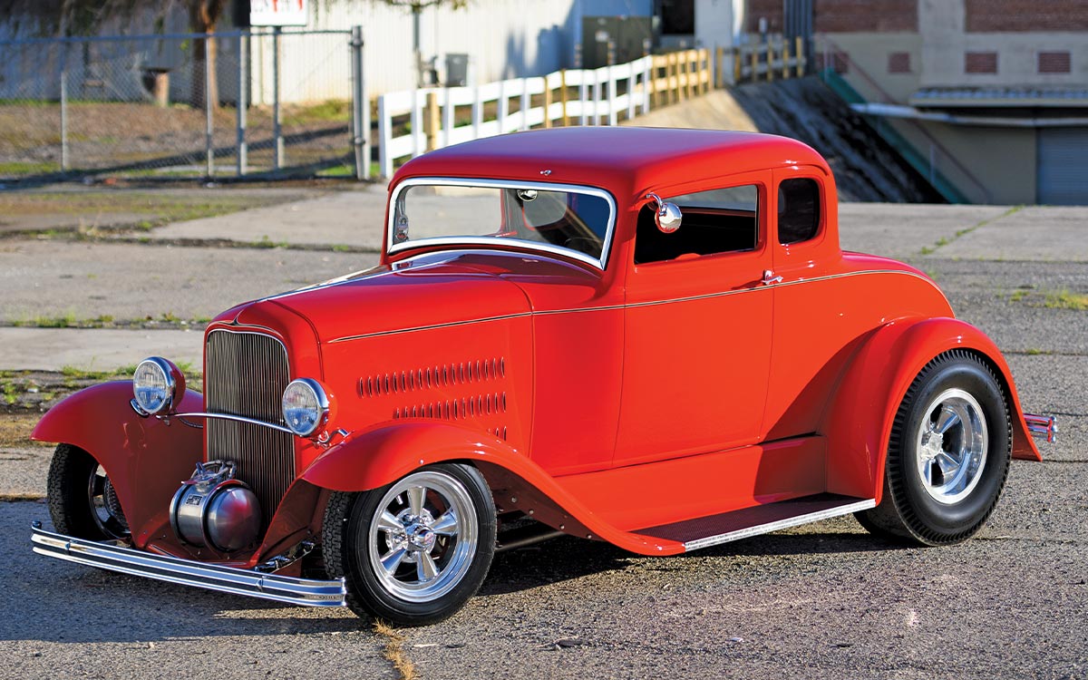 High-angle shot of the red '31 Ford Coupe hot rod on asphalt. Highlights the chopped roofline, louvers on the hood side panels, and the contrast between the slim front tires and massive rear racing tires on chrome five-spoke wheels.