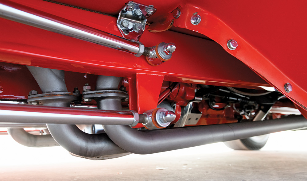 Undercarriage detail of the red '31 Ford showing the painted frame rail, chrome suspension links with acorn nuts, and matte gray ceramic-coated exhaust headers exiting from the engine block.