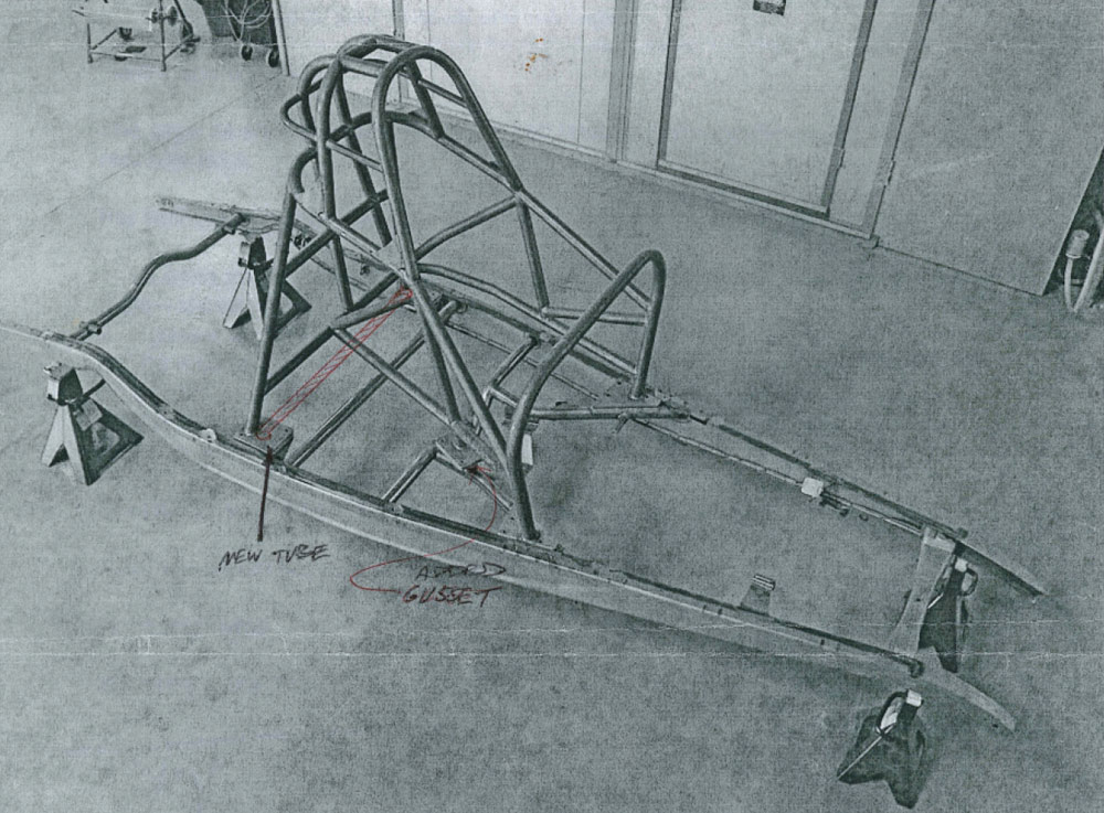 Grayscale photo of a Funny Car-style rollcage bolted to a car frame on jack stands. Red handwritten notes and arrows highlight modifications, specifically a "new tube" and an "added gusset" within the tubular steel structure.