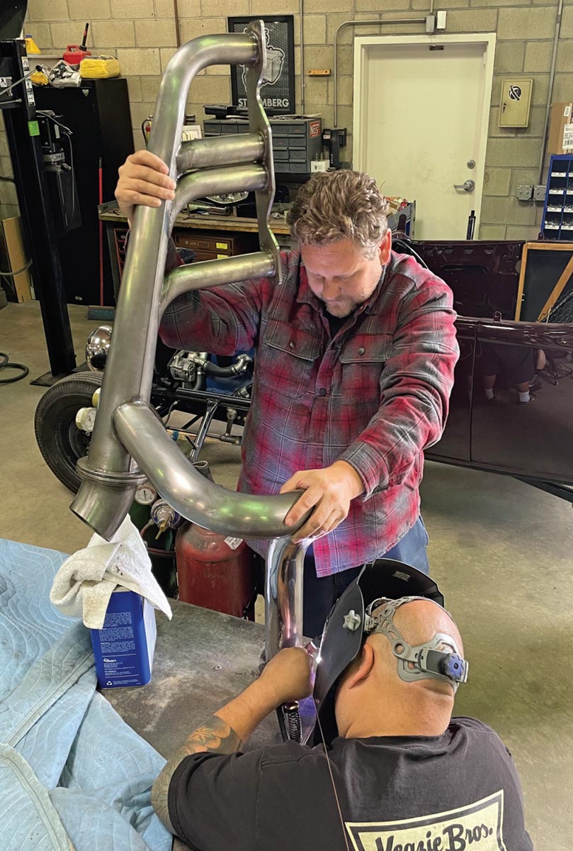 In a workshop, Evin Veazie holds a large, raw metal exhaust header vertically while Greg Hirota, wearing a welding helmet, TIG welds the bottom flange on a workbench.