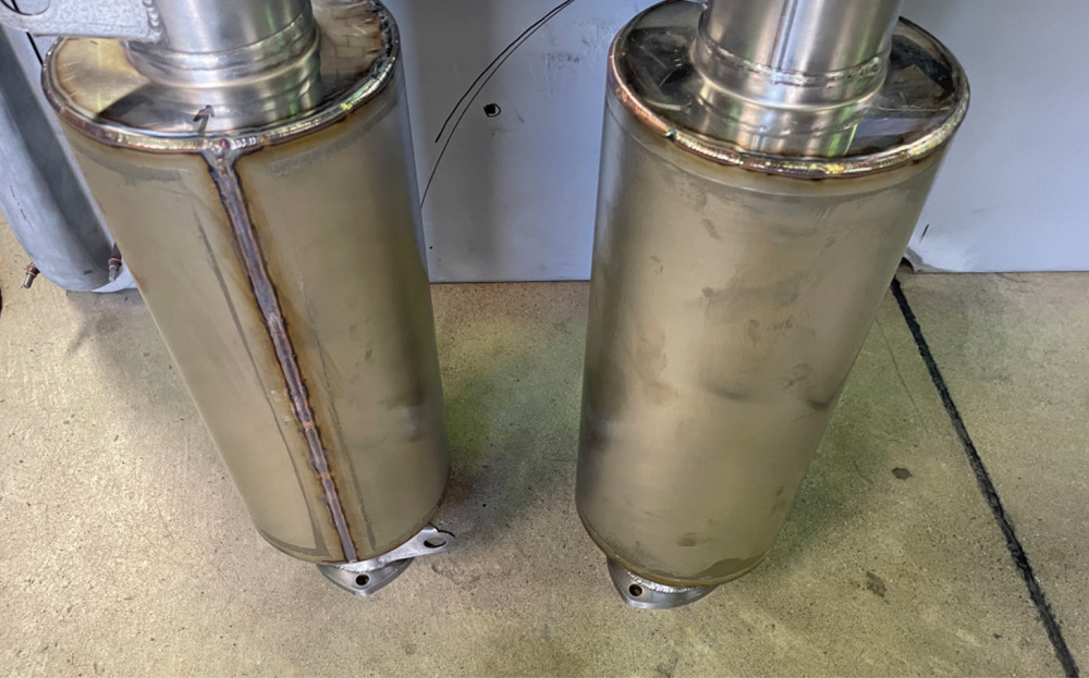 Top-down view of two cylindrical MagnaFlow mufflers. Each is encased in a smooth, polished 18-gauge stainless steel wrap with visible vertical weld seams, effectively concealing any original manufacturer branding.