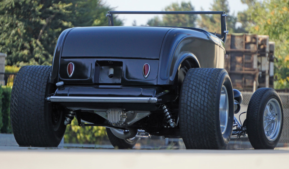 Low-angle rear view of the black 1932 Ford roadster in matte sealer. Features include oversized rear tires, teardrop taillights, and visible silver rear-end components and suspension under the chassis.