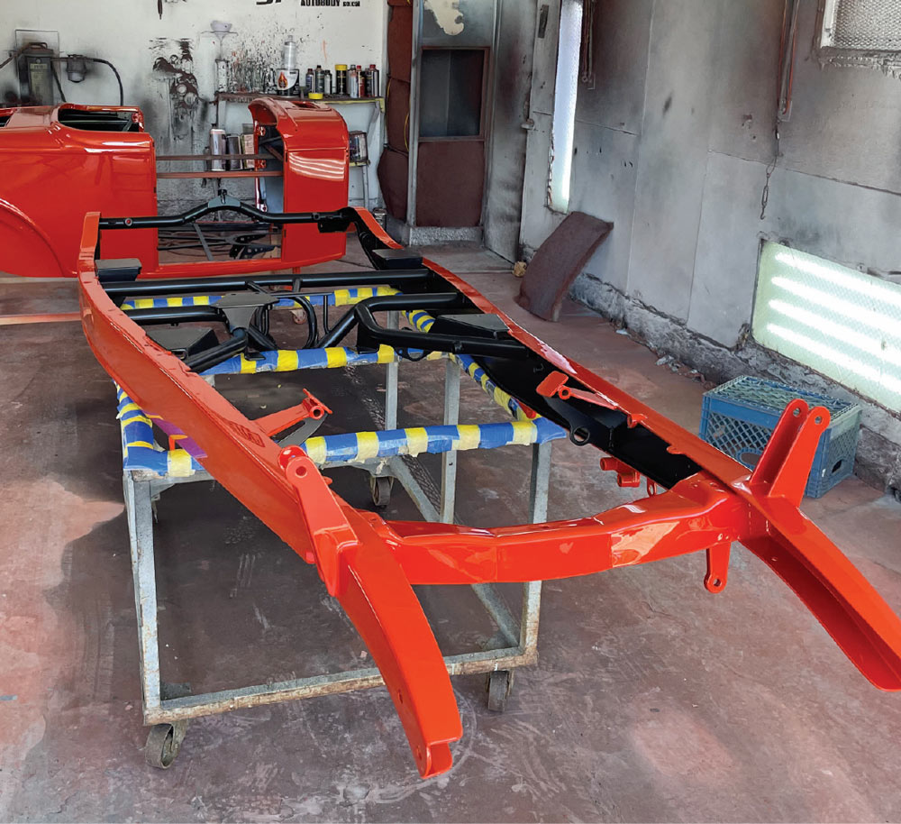 The roadster frame rests on a metal stand in a workshop. The outer perimeter rails are painted a vibrant burnt orange, while the internal crossmembers and boxed plates are finished in contrasting satin black.