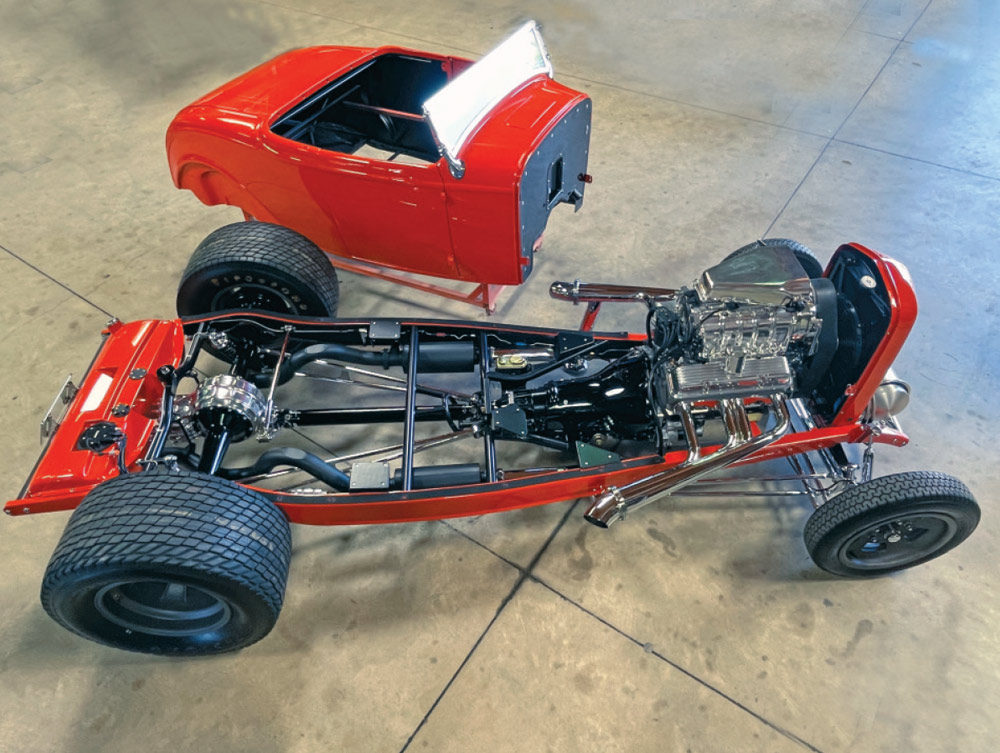 High-angle view of the red 1932 Ford body sitting beside its rolling chassis. The chassis displays orange frame rails, a supercharged engine, and the black TH400 transmission with a visible scattershield before the body is installed.