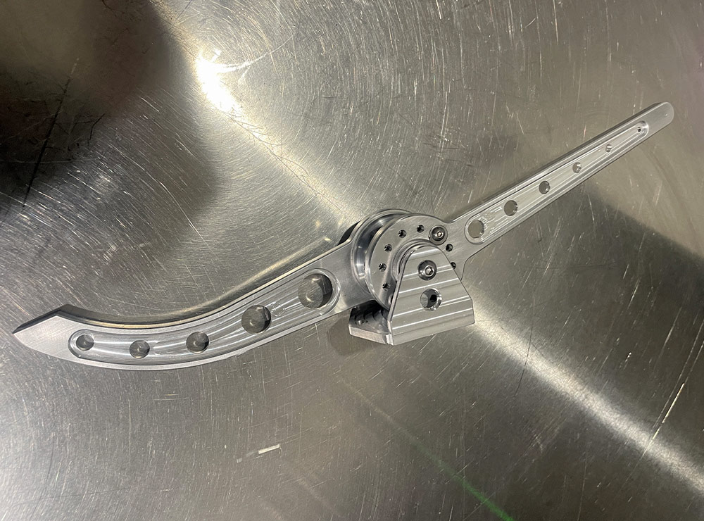 Machined aluminum throttle pedal assembly on a metallic surface. It features a curved arm with four circular lightening holes and a detailed central pivot bracket held by hex-head bolts.